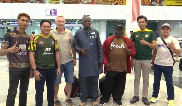 Seven men standing together in the arrivals hall at Kuala Lumpur International Airport, among them Tibor Szaban in a grey polo shirt, Olubunmi Adigun in a cream robe at the centre, and Peter Nieketien in a brown shirt, with officials in Kedah green alongside them